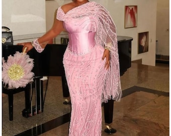 May include: A floor-length light pink gown featuring a corset-style bodice, intricate beadwork, and sequin embroidery. The dress includes a draped off-the-shoulder sleeve, a matching headwrap, and a coordinating feathered hand fan with a gold center.