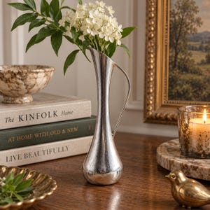 May include: A tall, slender silver-toned metal vase with a curved handle and a flared rim, containing a bouquet of small white flowers with green leaves. The vase has a smooth, reflective surface and a teardrop-shaped base, standing upright.