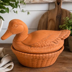 May include: A terracotta duck-shaped lidded container featuring a detailed molded duck figure on top of a round base with a textured basket-weave pattern. The entire piece is crafted from unglazed reddish-brown clay with visible feather details.