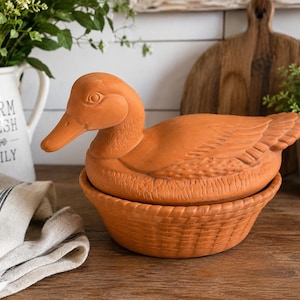 May include: A terracotta duck-shaped container with a basket-weave base. The duck has detailed feather and eye features. A white pitcher with the words "FARM FRESH DAILY" is in the background.