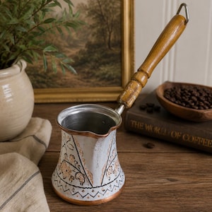 May include: A metal coffee pot with a white exterior featuring black and copper-colored floral patterns, a wide base, a pouring spout, and a long, cylindrical wooden handle with a metal loop at the end for hanging or storage.