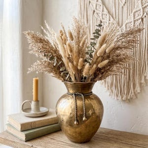 May include: A golden, hammered-metal vase filled with dried pampas grass, wheat, and eucalyptus. The vase has a rope detail around the neck and sits on a wooden table with books and a candle. A macrame wall hanging is in the background.