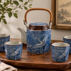 May include: A ceramic tea set featuring a hexagonal teapot with a woven handle and four matching cups. The pieces are decorated with a blue and white pattern depicting cranes flying over stylized waves, finished with a speckled off-white glaze.