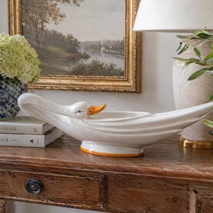 May include: A white ceramic swan-shaped serving dish with an orange beak and base trim. The dish is on a wooden table, with a painting and a lamp in the background. The swan's neck curves gracefully.
