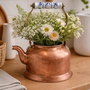 May include: A copper tea kettle filled with a bouquet of white and yellow flowers. The kettle has a white handle with blue floral designs. The flowers include daisies and other small white blossoms. The kettle sits on a wooden countertop.