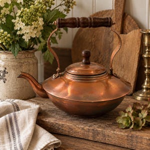 May include: A copper tea kettle with a wide, rounded base, a curved spout, and a removable lid featuring a dark brown wooden knob. The kettle has a tall, arched metal handle with a dark brown wooden grip, showcasing a polished metallic finish.