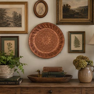May include: A round copper wall plate featuring an intricate embossed star and floral pattern with a textured border, displayed on a wall surrounded by framed botanical prints and landscape art above a wooden console table with decor.