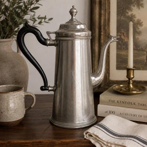 May include: A tall, metallic silver coffee pot with a smooth, cylindrical body, a domed lid featuring a small finial, and a curved black handle. The spout is elongated and slender, extending from the side of the vessel, which rests on a circular base.