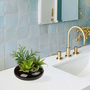 May include: A low, round, glossy black ceramic planter containing a cluster of green artificial succulents. The planter is positioned on a white countertop next to a gold-toned faucet and a set of light blue square wall tiles.