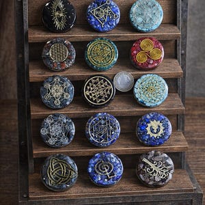 Op de afbeelding: Een houten displayplank bevat een verzameling ronde, decoratieve stenen. Elke steen heeft een uniek ontwerp met goudkleurige symbolen en patronen. De stenen zijn in verschillende kleuren, waaronder blauw, zwart, rood en grijs. De plank is gemaakt van donker hout.