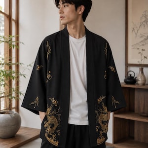 Könnte beinhalten: Eine schwarze, vorne offene Jacke im Kimono-Stil mit goldfarbenen Drachenstickereien an den unteren Vorderteilen und geometrischen Linienmustern an den Ärmeln. Das Kleidungsstück wird über einem schlichten weißen T-Shirt und einer schwarzen Hose getragen.