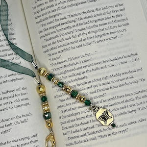 May include: A bookmark featuring a dark green ribbon attached to a series of gold-toned metal beads and faceted green glass beads. The end of the bookmark displays a gold-toned metal charm shaped like a butterfly with intricate wing details.