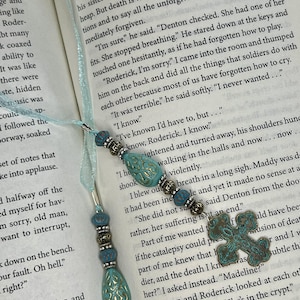 May include: A bookmark featuring a light blue ribbon with two dangling beaded strands. Each strand includes turquoise-colored teardrop beads with gold-toned filigree patterns, silver-toned spacers, and a metal cross charm with a weathered patina.