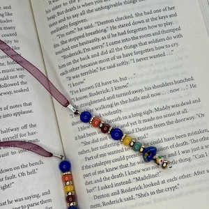 May include: Two beaded bookmarks resting on an open book page. Each bookmark features a dark purple ribbon attached to a silver metal loop, followed by a sequence of blue, orange, yellow, red, and teal beads, ending with a large blue bead.