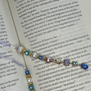 May include: Two beaded bookmarks resting on an open book page. Each bookmark features a light purple ribbon attached to a series of beads, including white pearls, faceted blue crystals, and smooth lavender stones, separated by gold-toned metal spacers.