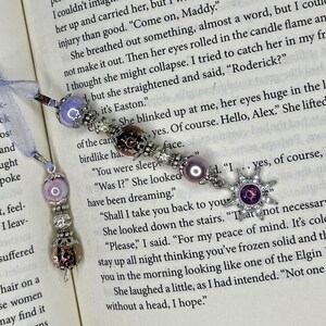 May include: Two beaded bookmarks with decorative charms. One features a purple bead and a silver charm, the other has a starburst charm with a purple center. Both are on a book page with text.