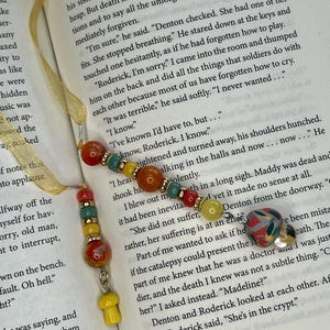 May include: Two beaded bookmarks featuring gold-toned ribbons and assorted beads in red, orange, yellow, and teal. One bookmark includes a small yellow mushroom charm, while the other features a multi-colored patterned mushroom charm.