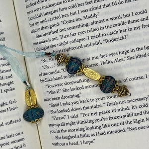 May include: Two decorative bookmarks with blue and gold accents rest on an open book. One bookmark features a gold oval pendant with a blue bead, and a light blue ribbon. The other has a series of blue and gold beads.
