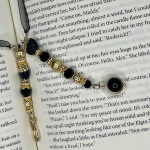 May include: Elegant black and gold beaded bookmark with a black ribbon. The bookmark features decorative beads and a charm, resting on the pages of a book. A stylish accessory for any book lover.