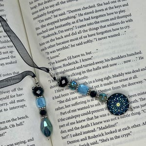 May include: Two bookmarks featuring black ribbons and beaded dangles. The beads include black, light blue, and teal faceted glass, with one bookmark ending in a round, dark blue glass charm featuring a yellow and white starburst pattern.