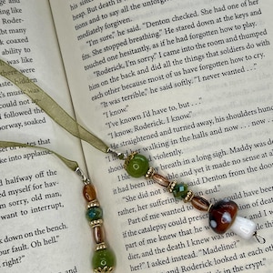 May include: Two bookmark charms featuring olive green ribbons and beaded dangles. The beads include round green stones, faceted amber and green glass, gold-toned spacers, and a small brown and white mushroom-shaped glass bead at the end.