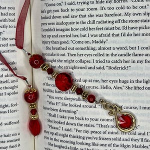 May include: A red beaded bookmark with a gold-colored charm and a red ribbon. The bookmark features various red beads and a decorative charm with a red gem. The bookmark is placed in an open book.
