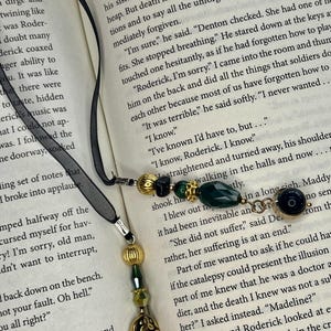 May include: A black ribbon bookmark featuring two dangling ends adorned with gold-toned metal beads, dark green faceted glass beads, and a small oval gold-toned charm with an intricate embossed pattern, resting on an open book page.