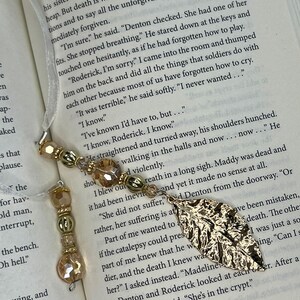 May include: A bookmark featuring a gold-toned metal leaf charm attached to a white ribbon. The ribbon is adorned with a series of light brown, translucent, and gold-colored beads in various shapes, including faceted spheres and metallic spacers.