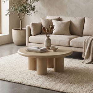 Puede incluir: Una mesa de centro redonda de madera clara con tres patas cilíndricas. La mesa está frente a un sofá beige con cojines a juego. Un pequeño jarrón con flores secas y un cuenco están sobre la mesa. Una alfombra crema está en el suelo.