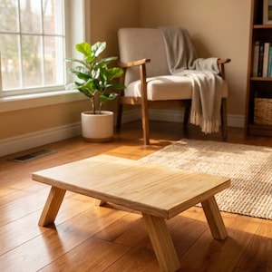 May include: A small, rectangular wooden table with angled legs. The table is made of light-colored wood and is positioned on a hardwood floor. The room features a window, a chair, and a potted plant, suggesting a living room setting.