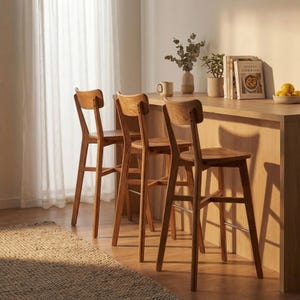 Op de afbeelding: Drie houten barkrukken met gebogen rugleuningen en slanke poten zijn geplaatst voor een lichtgekleurde houten toonbank. De krukken hebben een natuurlijke houten afwerking. Een tapijt bevindt zich op de voorgrond en een transparant gordijn op de achtergrond.