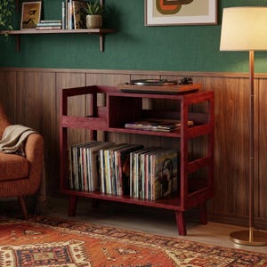 Puede incluir: Un soporte de madera rojo para tocadiscos con un estante incorporado para discos de vinilo. El soporte tiene un tocadiscos en la parte superior y está lleno de discos de vinilo. El soporte está en una habitación con una alfombra y una lámpara.