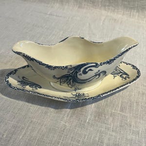 May include: A cream-coloured ceramic gravy boat with an attached oval underplate, featuring a dark blue floral and scroll pattern along the rim and sides. The piece has a curved, elongated spout and a handle on the opposite side, with a smooth finish.