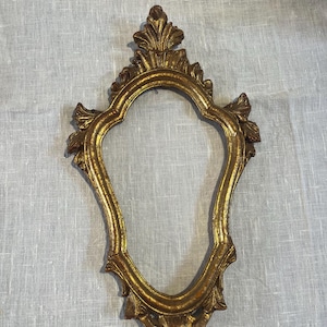 May include: An ornate, gold-finished picture frame with a distinct, elongated, asymmetrical shape. The frame features intricate, carved leaf-like detailing at the top and sides, with a weathered, antique texture throughout its gilded surface.