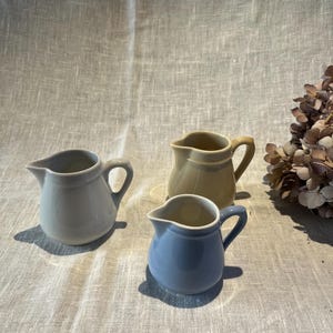 Può includere: Tre piccole lattiere in ceramica nei colori grigio chiaro, giallo pallido e blu tenue. Ogni brocca presenta un corpo arrotondato, un manico curvo e un beccuccio distinto. I recipienti sono disposti su una superficie in tessuto testurizzato vicino a fiori secchi.