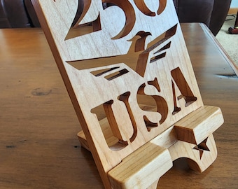Puede incluir: Un soporte de teléfono de madera con las palabras "FREEDOM 250 USA" recortadas. El soporte presenta un diseño de águila y recortes de estrellas. La madera es de color marrón claro y está inclinada para su exhibición.