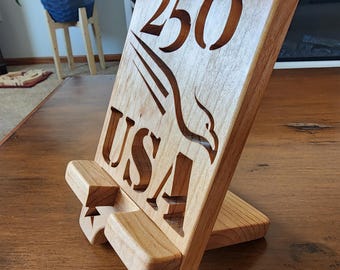 Puede incluir: Un soporte de tableta de madera clara con el texto "1776-2026 250 USA" y un diseño de águila. El soporte tiene una ranura para sostener una tableta o un libro y está inclinado para facilitar la visualización.