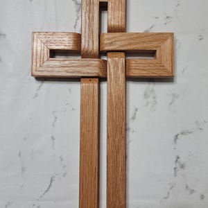 May include: A wooden cross with a unique, interwoven design. The cross is constructed from light brown wood, featuring rectangular frames that intersect to form the traditional Christian symbol. The wood grain is visible, adding texture to the piece.