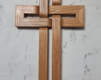 Puede incluir: Una cruz de madera con un diseño único e entrelazado. La cruz está hecha de madera marrón claro, con marcos rectangulares que se cruzan para formar el símbolo cristiano tradicional. La veta de la madera es visible, agregando textura.