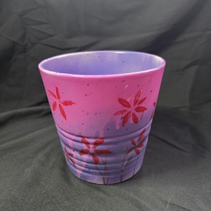 May include: A hand-painted ceramic flower pot with a gradient of pink and purple. Red flower designs are painted around the pot. The pot has a slightly flared shape and is suitable for indoor or outdoor use.