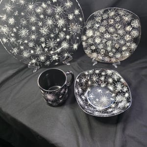 Op de afbeelding: Een zwart-wit servies bestaande uit een groot rond bord, een vierkant bord, een mok en een kom. Elk stuk is versierd met een wit bloemmotief. De set wordt gepresenteerd op een zwart oppervlak.