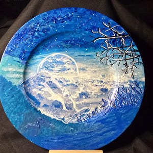 May include: A round decorative plate with a hand-painted landscape scene. The plate features a blue and white colour palette, depicting a moonlit mountain scene with a starry sky and a bare tree branch.