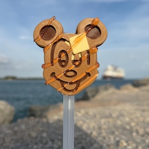May include: A Mickey Mouse-shaped waffle on a stick, drizzled with caramel and topped with a pat of butter. The treat is brown with dark brown accents and sits against a blurred background of water and sky.