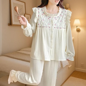 May include: Cream-colored pajama set with a long-sleeved top and pants. The top features a ruffled collar and floral embroidery. The fabric has a ribbed texture. The set includes a button-down top and matching pants, creating a comfortable and coordinated sleepwear ensemble.