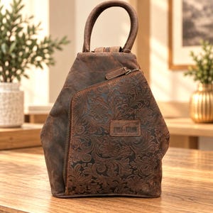 Könnte beinhalten: Ein brauner Lederrucksack mit Tragegriff und einer diagonalen Reißverschlusstasche auf der Vorderseite. Die Frontpartie zeigt ein geprägtes Blumenmuster und ein rechteckiges Etikett mit dem Text BLUE FURY. Der Rucksack ist konisch geformt.