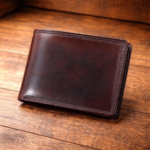 Op de afbeelding: Een donkerbruine lederen bifold portemonnee met een gladde, gepolijste afwerking en zichtbare ton-sur-ton stiksels langs de randen. De portemonnee heeft een rechthoekige vorm met afgeronde hoeken en een consistente, diepe mahoniekleur over het oppervlak.