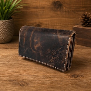 Peut inclure: Un portefeuille en cuir marron foncé avec une finition vieillie et un motif floral gaufré sur la surface avant. Le portefeuille présente des coutures de couleur claire visibles le long des bords et une construction multicouche pliée.