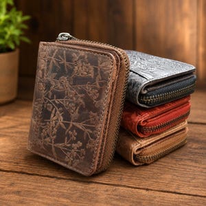 Peut inclure: Une pile de quatre petits portefeuilles zippés présentant un motif floral gaufré sur la surface. Les portefeuilles sont de couleur marron foncé, gris anthracite, rouge vif et beige clair, chacun doté d'une fermeture éclair en métal.