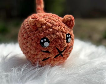 Peut inclure: Un petit jouet de chat orange en crochet avec des yeux noirs et un nez blanc. Le chat est assis sur une surface blanche et moelleuse.