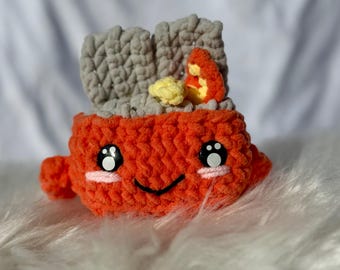 Peut inclure: Un rouleau de sushi au crochet orange fait à la main avec un visage souriant, des yeux noirs et des joues roses. Le sushi a du "riz" gris et une "garniture" jaune et orange. Le sushi est sur une surface blanche et moelleuse.